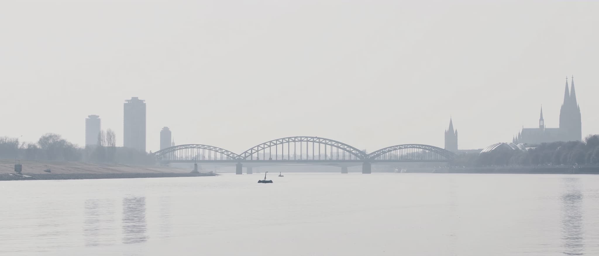 A steel truss bridge over the Rhine in haze, the towers of the city as shadows behind it