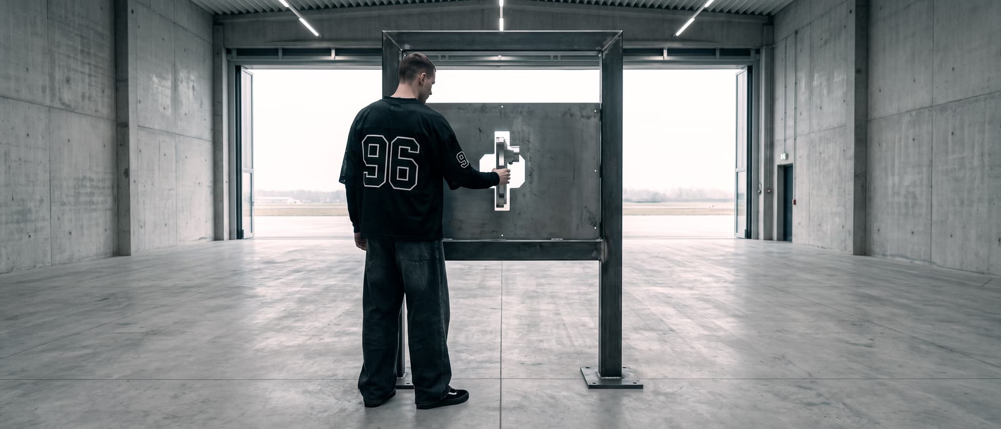 A steel inspection gate standing alone on the hangar floor, one plate with a precisely cut opening in it, a man seen from behind guiding a part into the cut